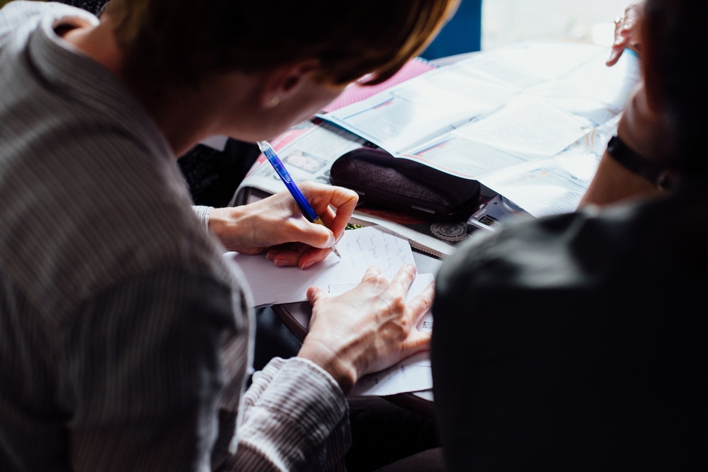Group of people sitting around table. One women is taking notes writing with pencil on paper.